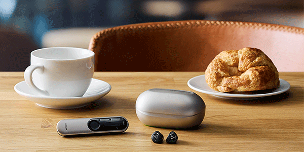 White cup and pastry on saucers beside earbuds, a small device, and pebbles on a wooden table against a brown chair.