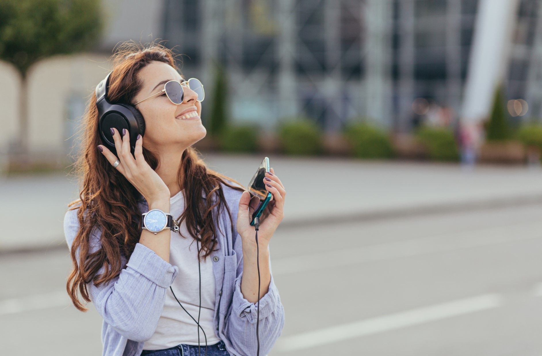 Woman wearing headphones and sunglasses walking outdoors while holding a phone and smiling