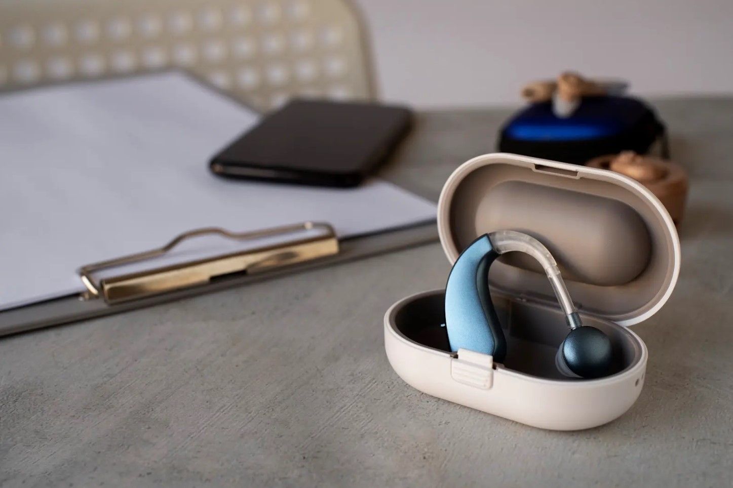 Open beige earbud case with blue and black earbuds on a desk, with a phone and pen in the background