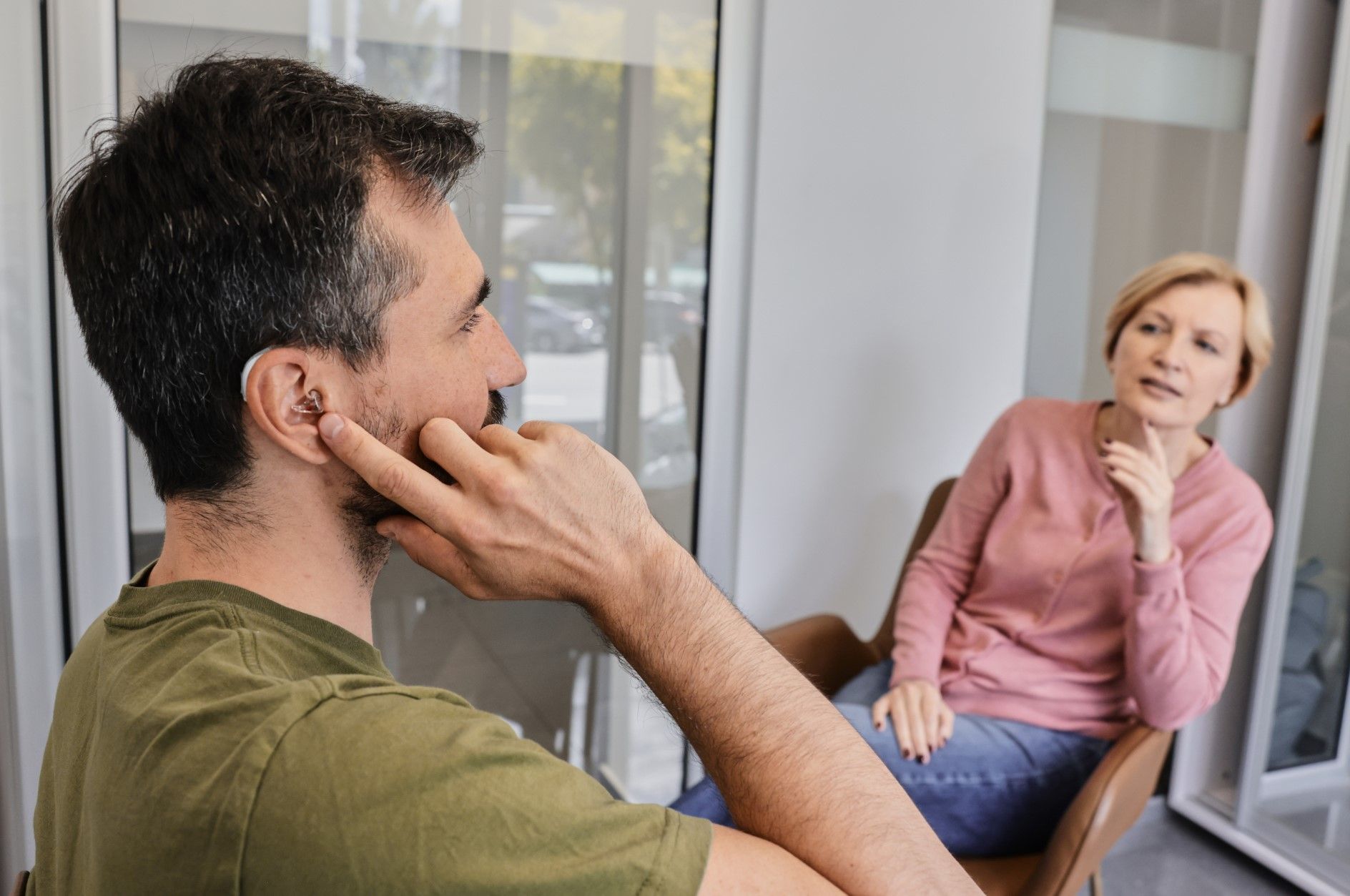 Two people talking by a window, one in a green shirt and one in a pink top seated on a chair.