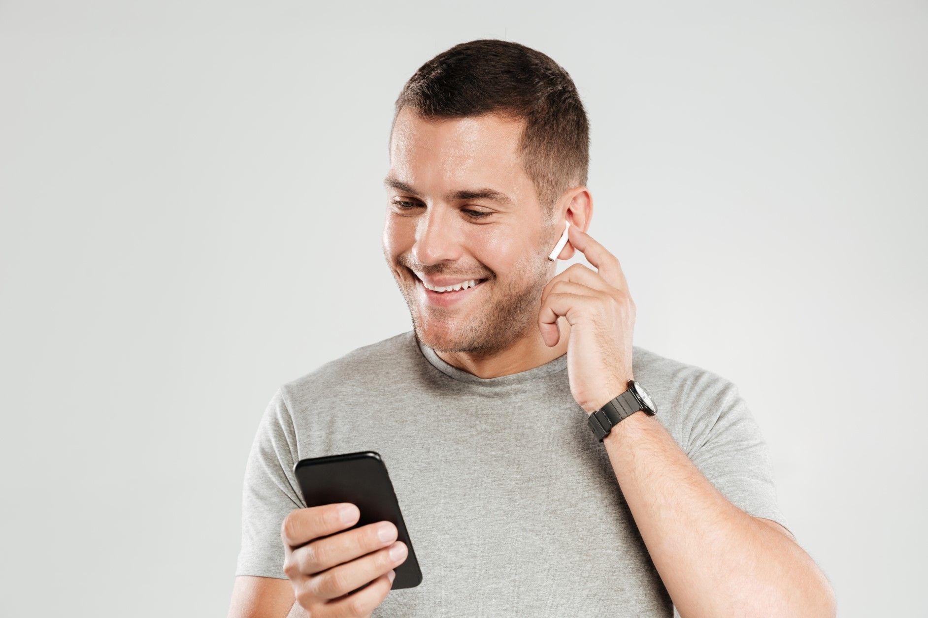 Smiling man in a gray shirt looking at a smartphone while adjusting a wireless earbud