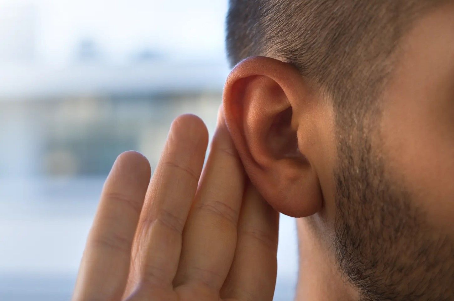 Hand cupped behind a man’s ear, listening intently outdoors with a blurred background
