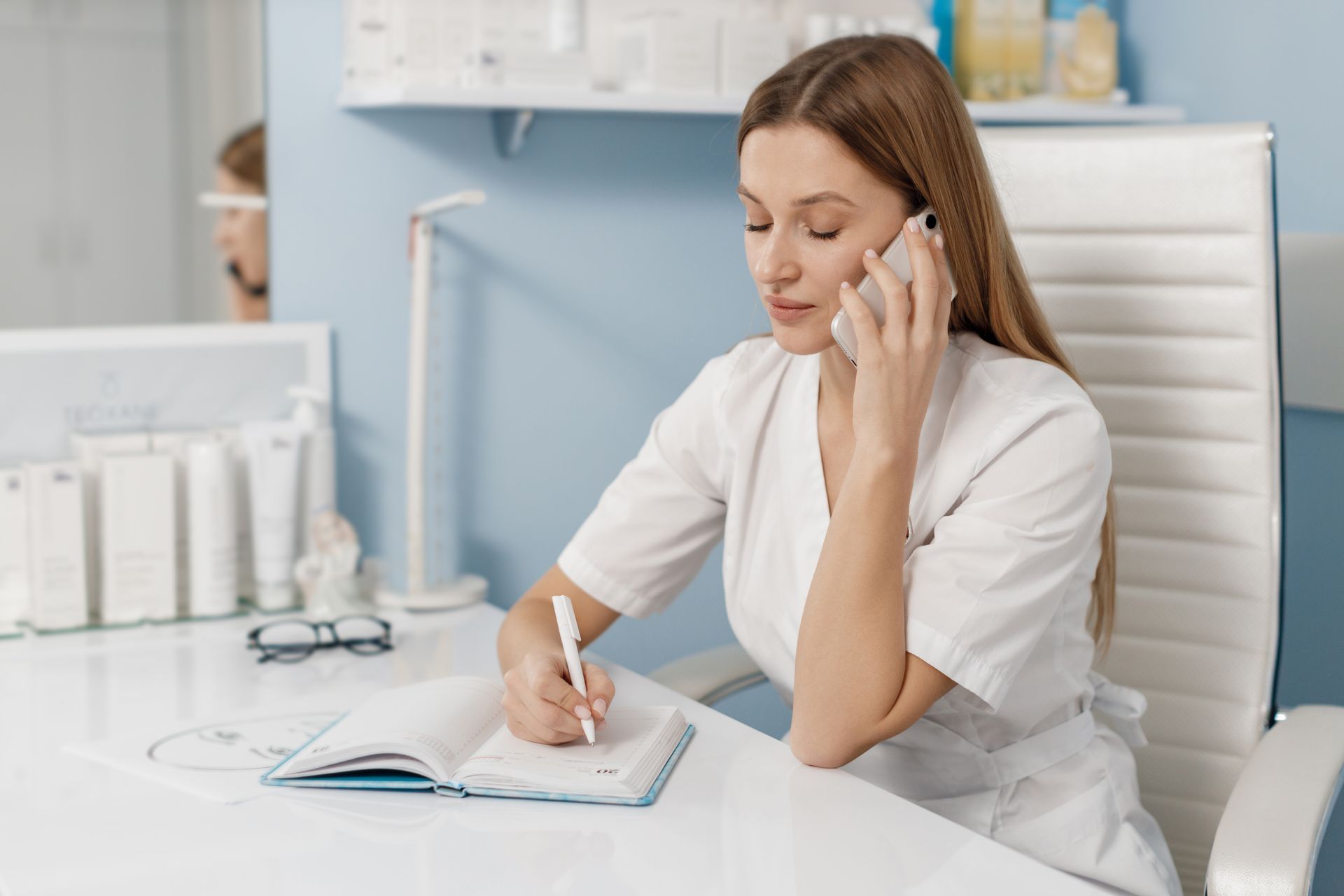Woman in white uniform writing in a notebook while talking on a phone in a bright office