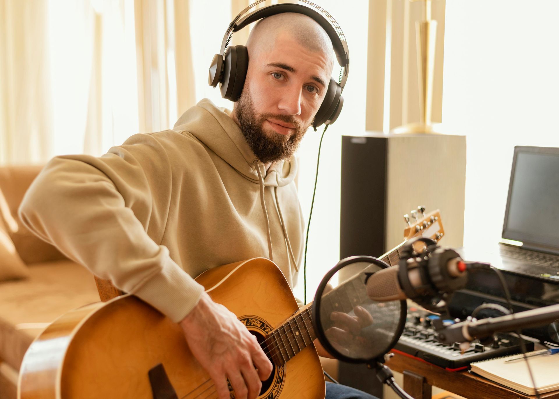 Man wearing headphones plays acoustic guitar beside a microphone and laptop in a home studio