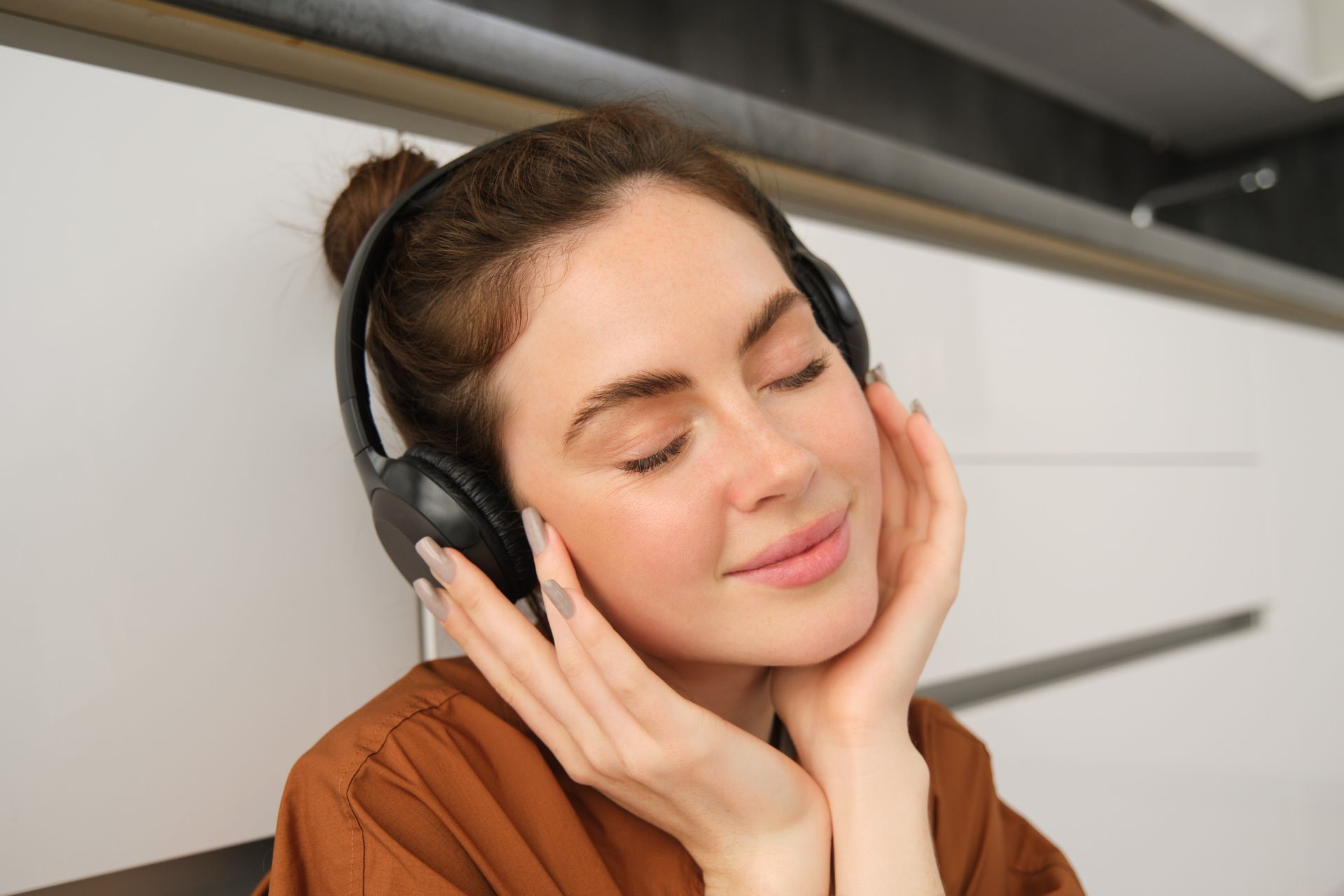 Woman wearing headphones, eyes closed, resting her face against her hands in a bright indoor setting