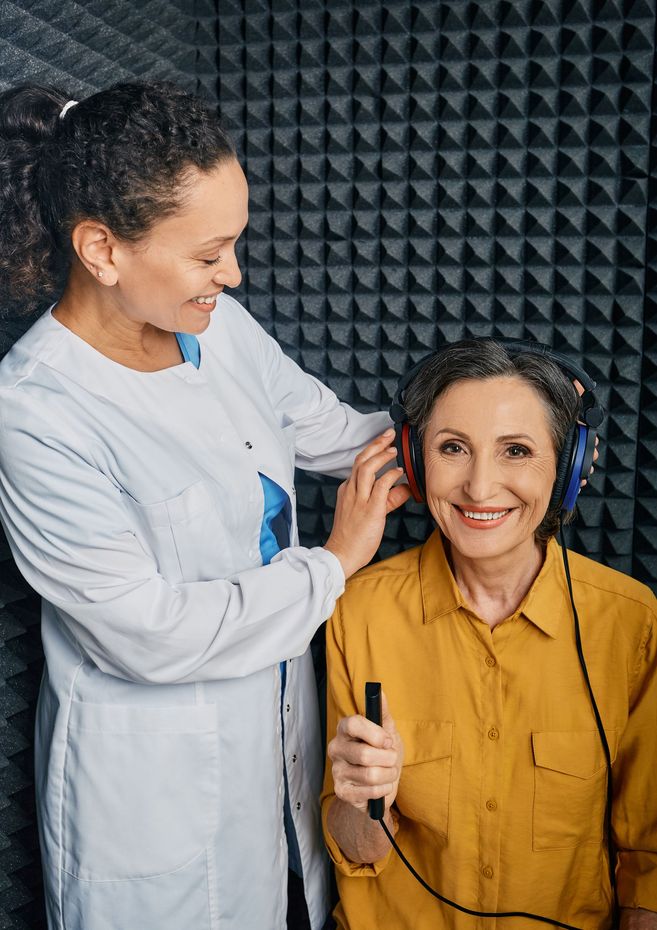 Two people in a soundproof studio, one adjusting the other’s earpiece as they smile at the camera.