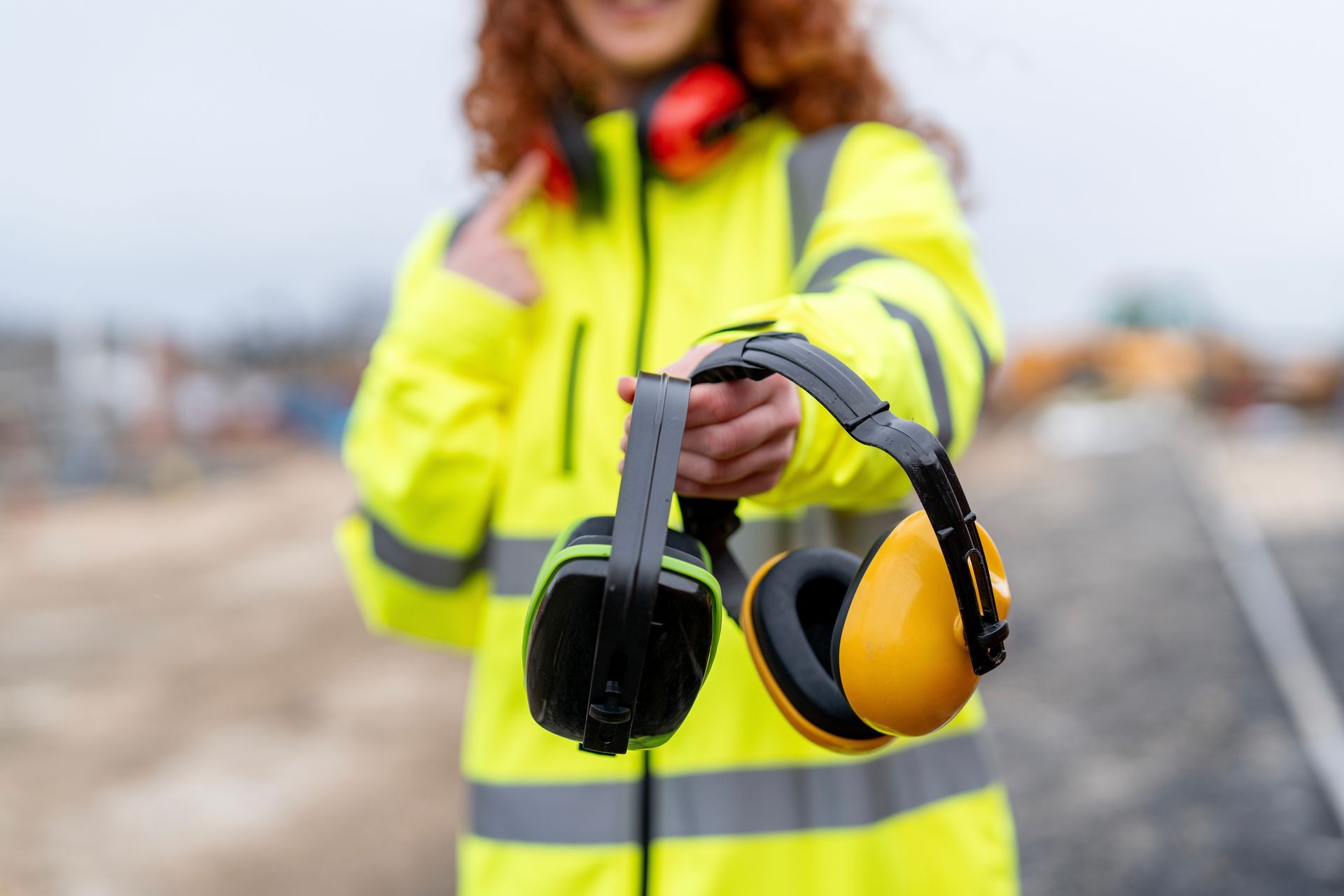 Person in a neon yellow safety jacket holding black-and-yellow headphones at a roadside construction site