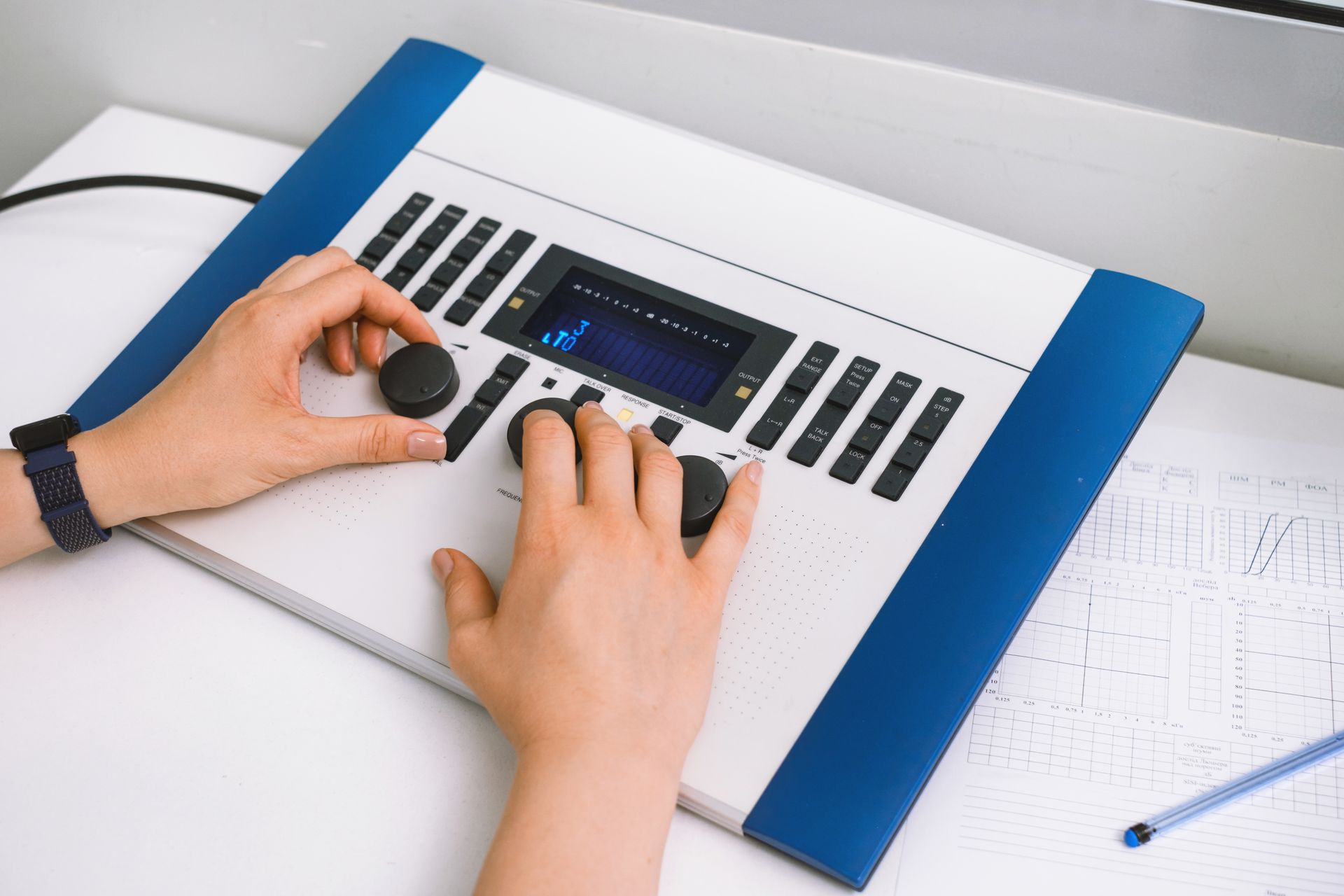 Hands adjusting controls on a white-and-blue audio mixer with a display and notebook nearby