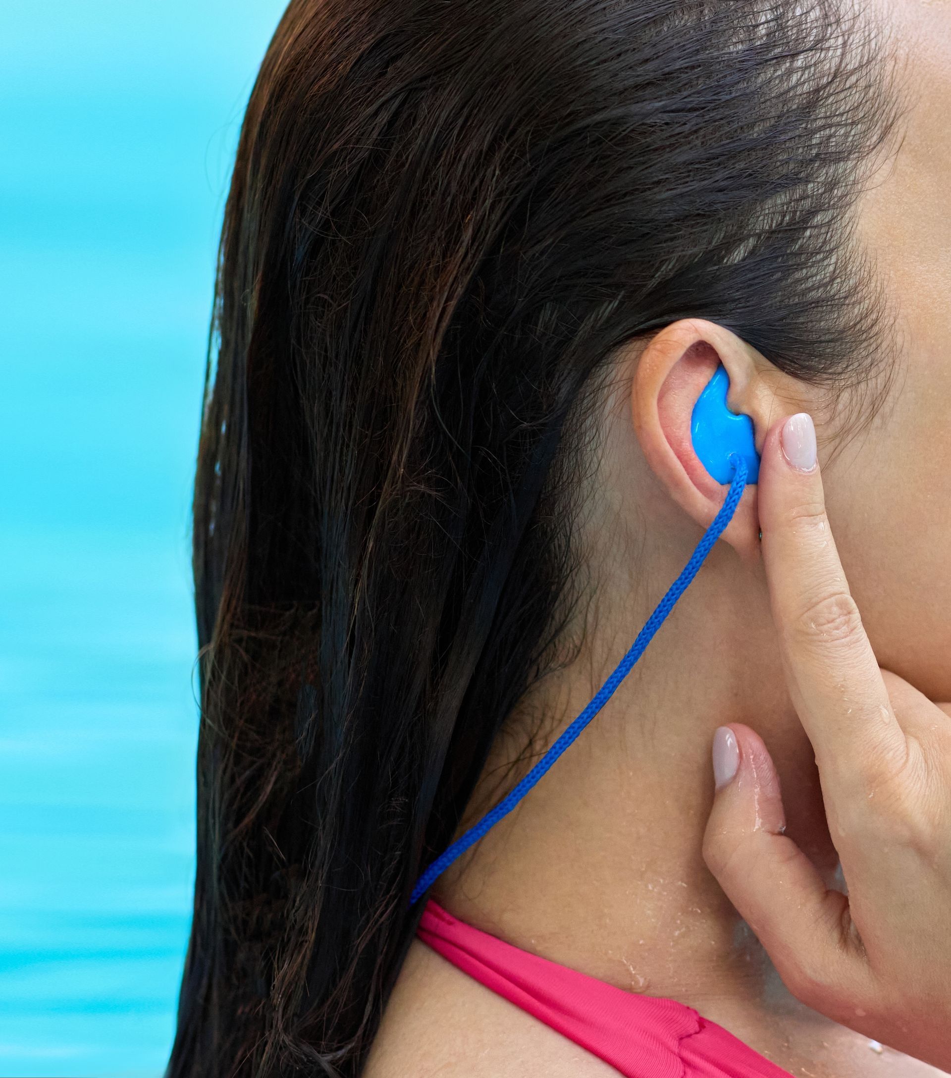 Woman wearing blue earplugs, holding one in place against a swimming pool background