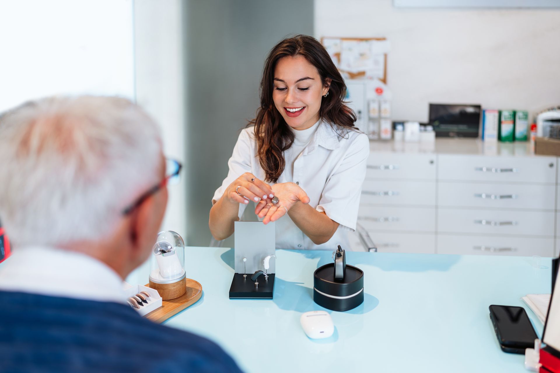 A support executive demonstrating a hearing aid model to a seated patient in a bright clinic room.