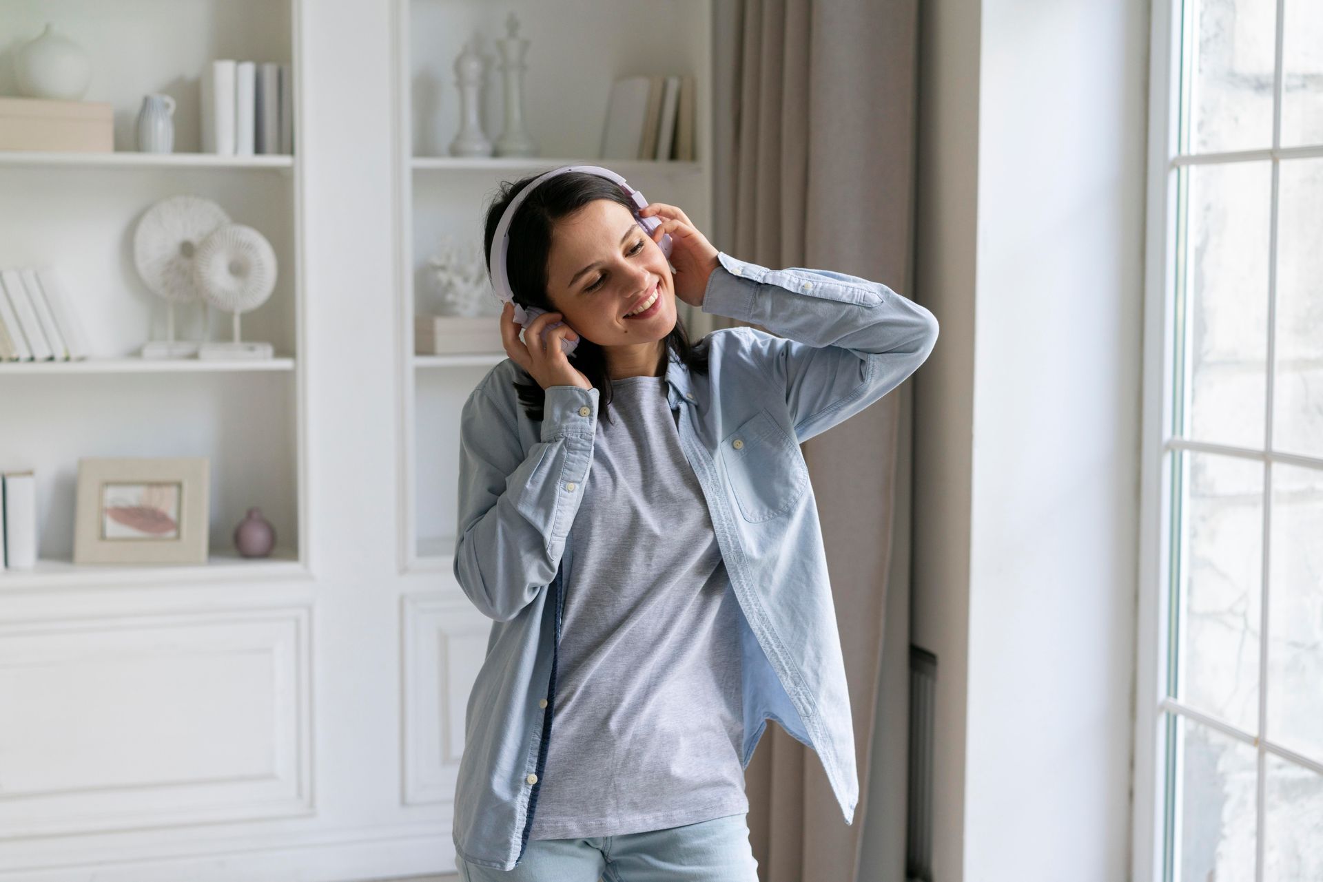 Person wearing headphones adjusts them while standing in a bright room by a window.