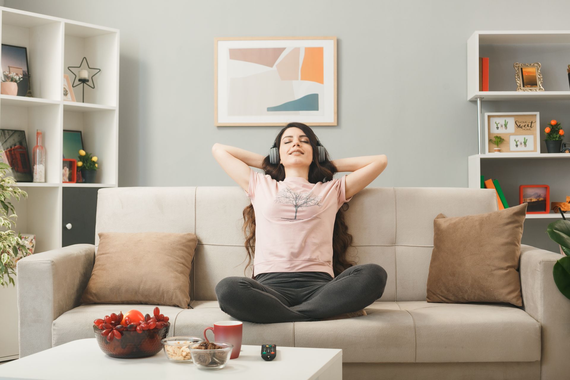 Person relaxing on a beige sofa in a bright living room, hands behind head, with shelves and wall art nearby