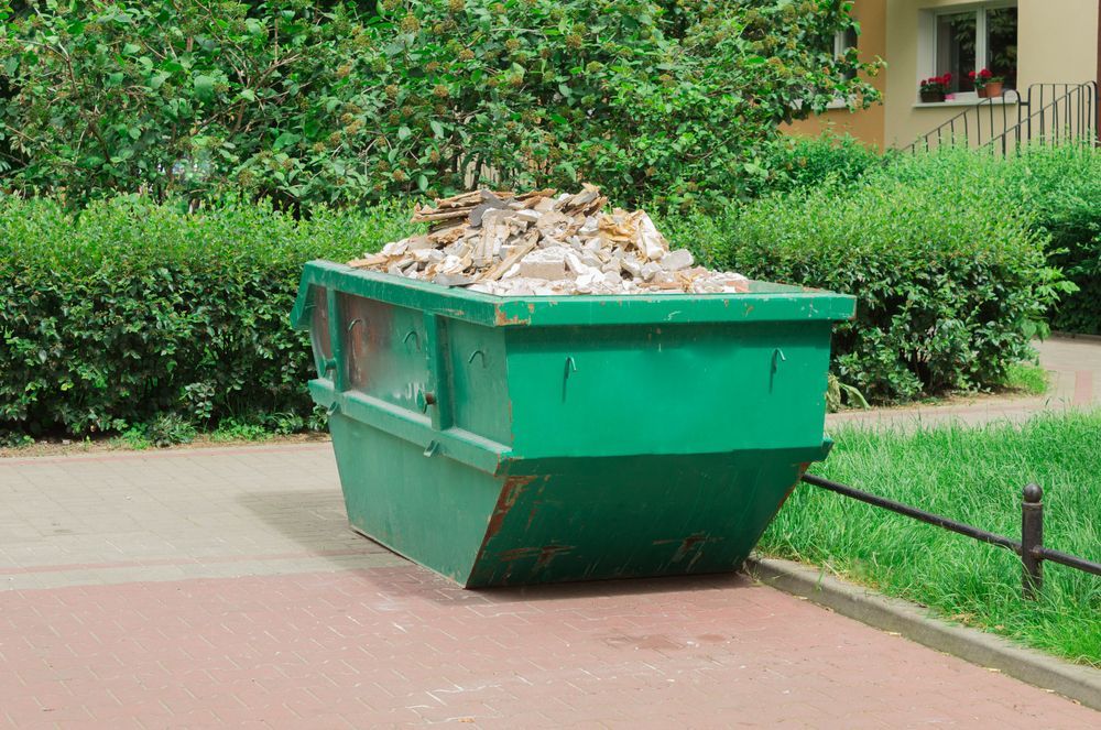 Green Dumpster Overflowing With Debris On A Sidewalk Next To A Hedge — Mount Isa Skips In Camooweal, QLD