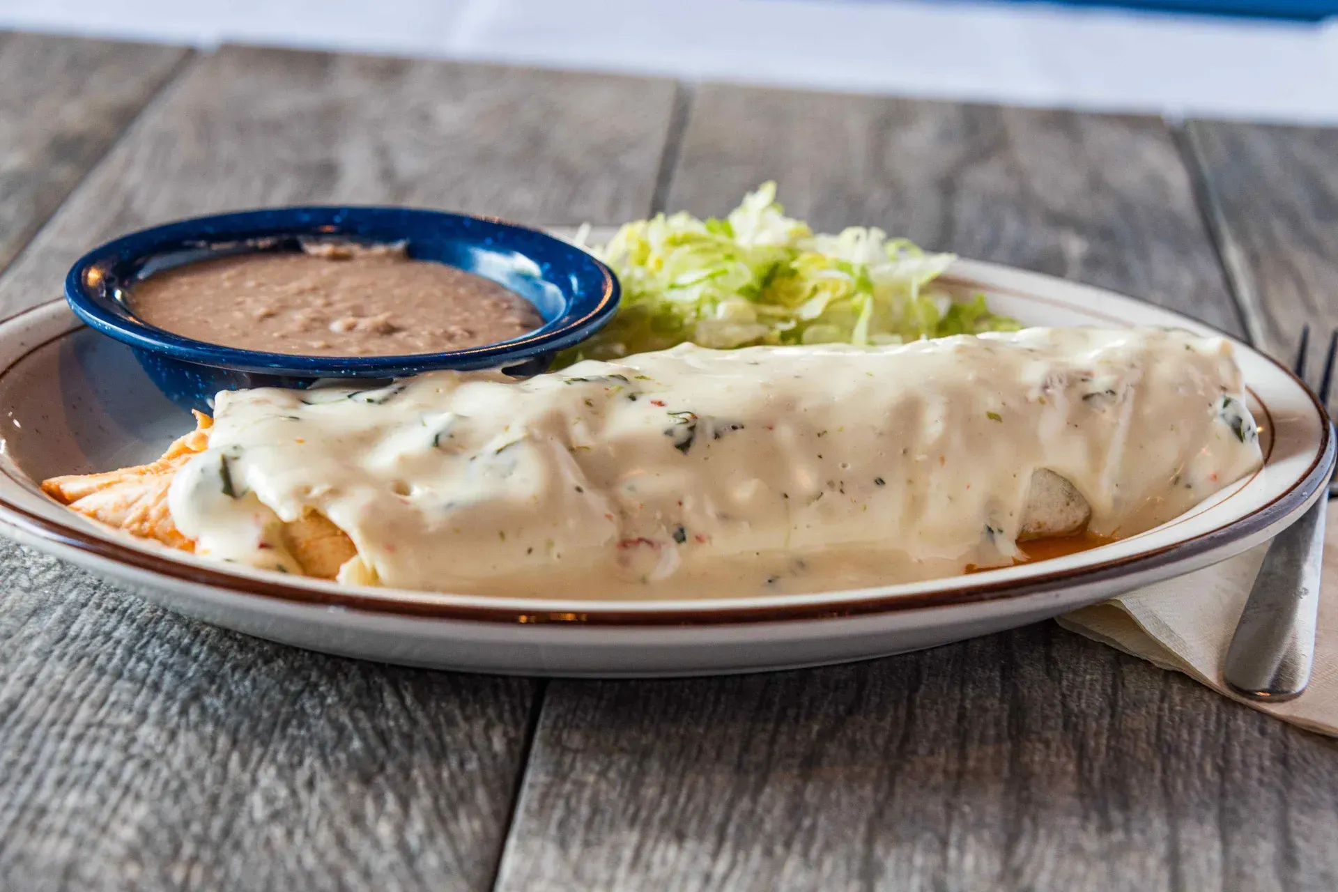 Enchilada with creamy white sauce, refried beans, and shredded lettuce on a plate.
