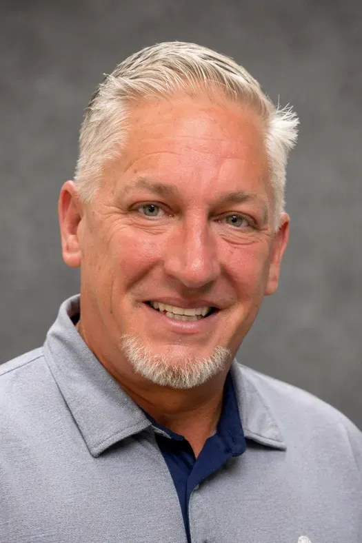 Smiling man with short gray hair wearing a light blue polo against a gray studio background