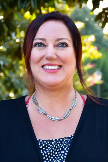 Smiling woman in a black top and silver necklace, standing outdoors with blurred trees behind her