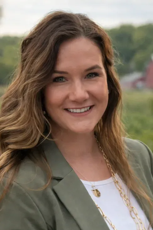 Smiling woman with long brown hair wearing a green blazer, standing outdoors with a blurred background.
