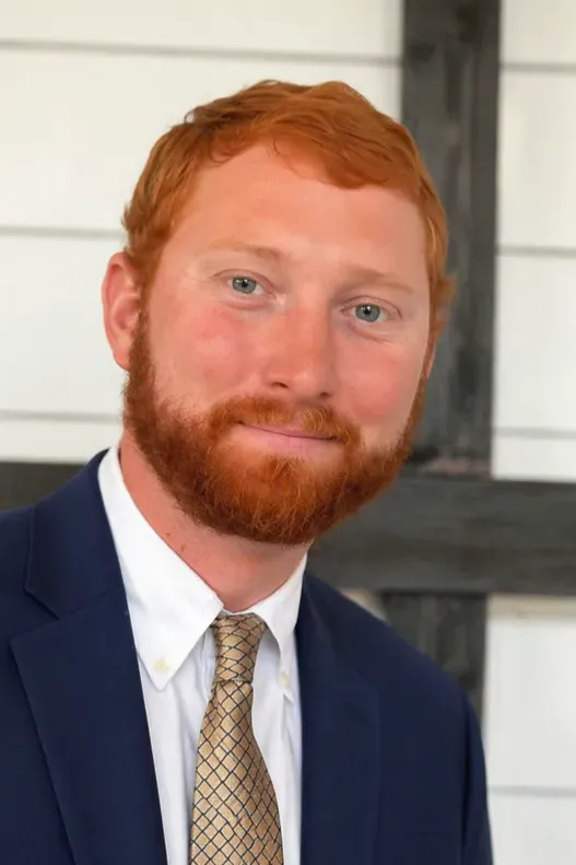 Portrait of a red-haired man in a navy suit and tie standing indoors against a white wall