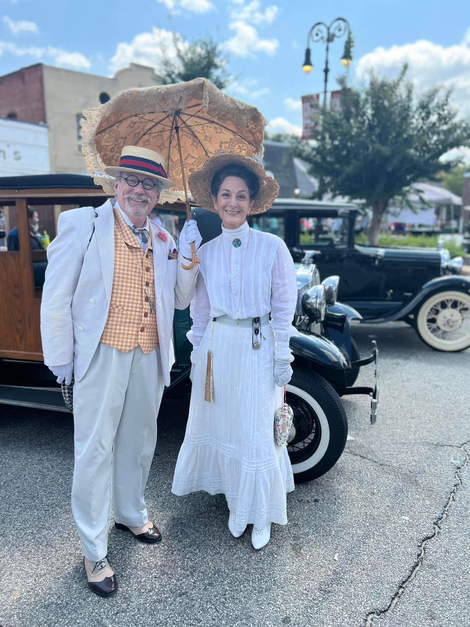 Two people in vintage-style outfits posing beside a classic car on a sunny street.
