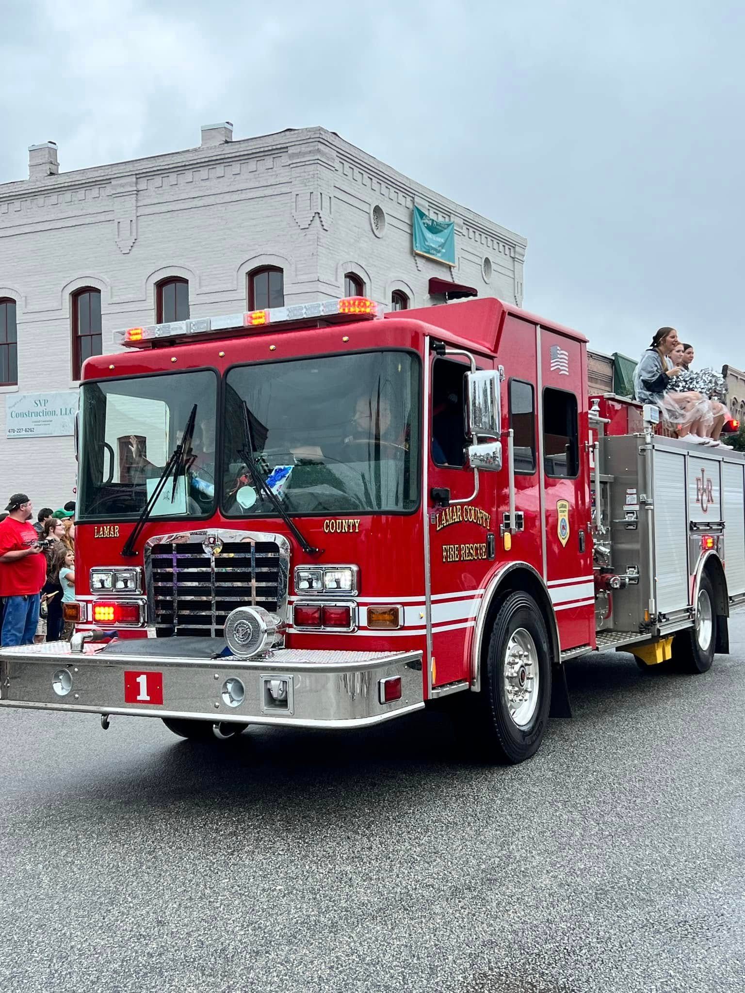 Red fire truck driving past a stone building, with people riding on the back.