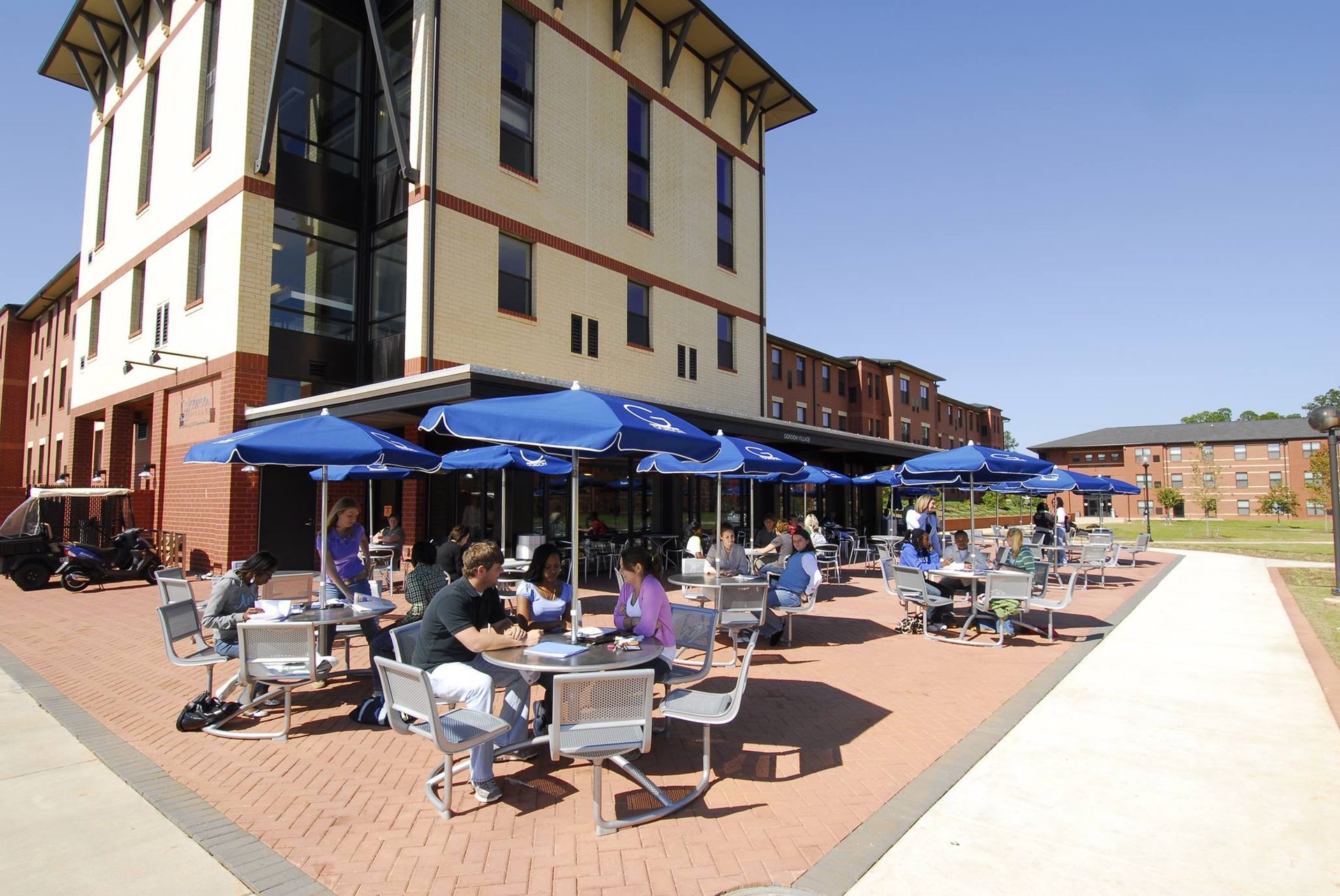 Outdoor café with people seated at blue umbrellas beside a tall building on a sunny day