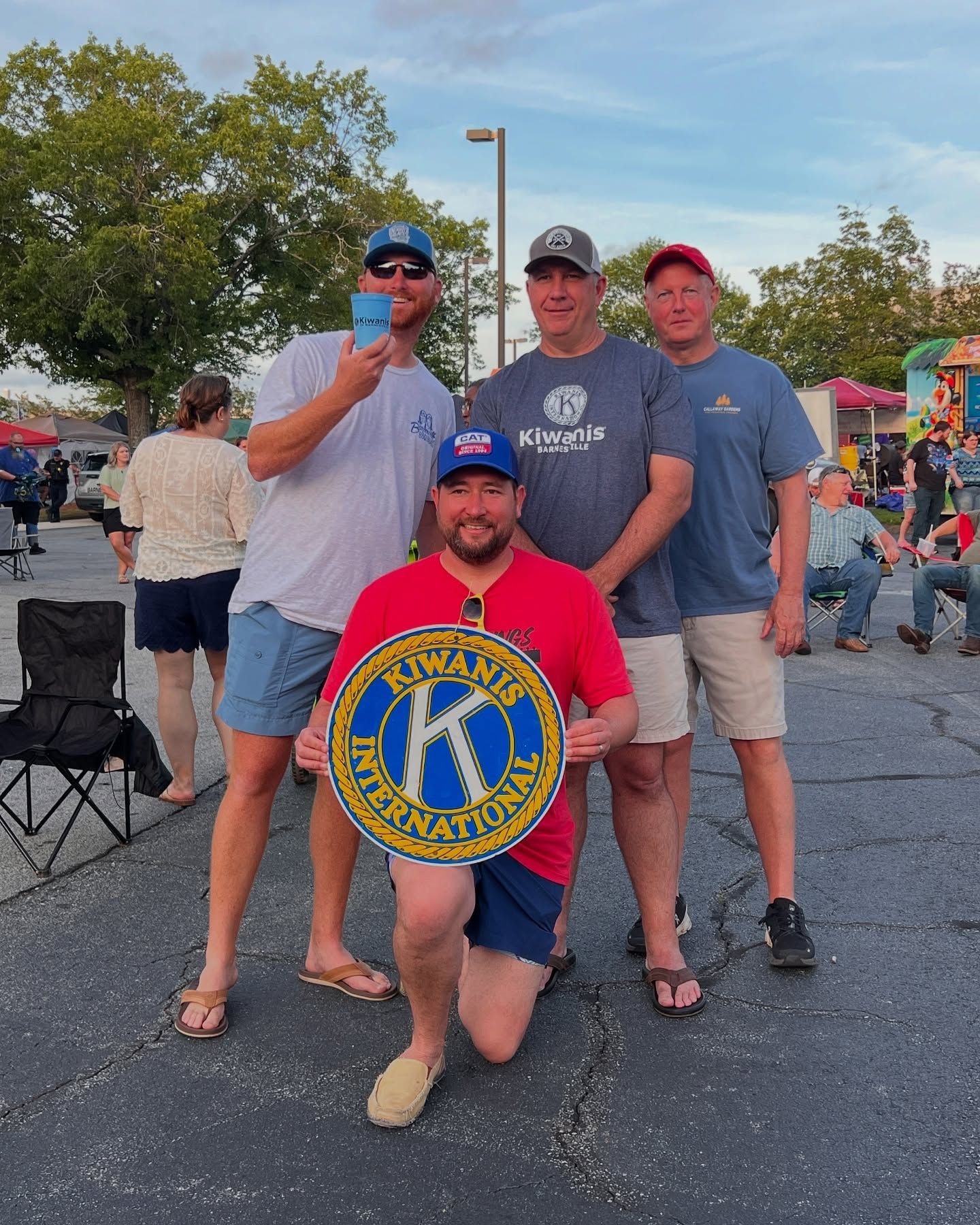 Four adults posing outdoors at a community event, one kneeling with a colorful circular sign.
