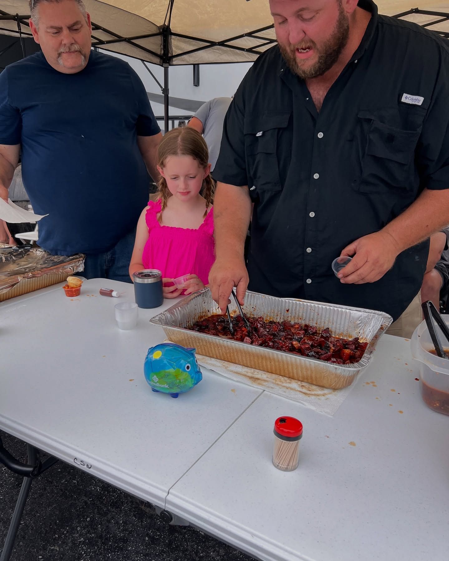 People gathered at a table outdoors, with a young girl and a large tray of food under a canopy.