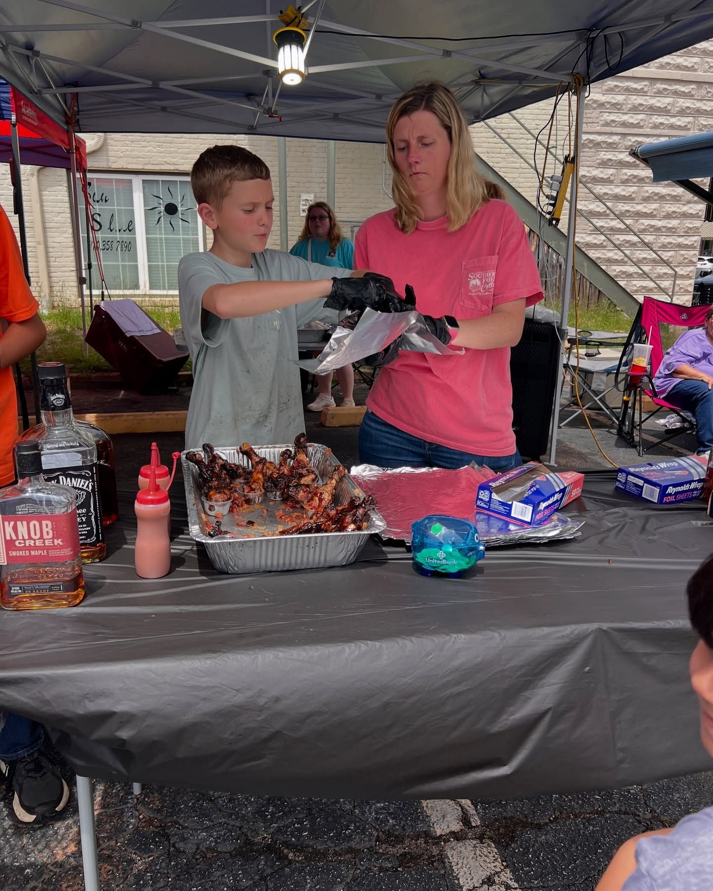 Two people serving food at an outdoor table under a canopy with trays, condiments, and drinks