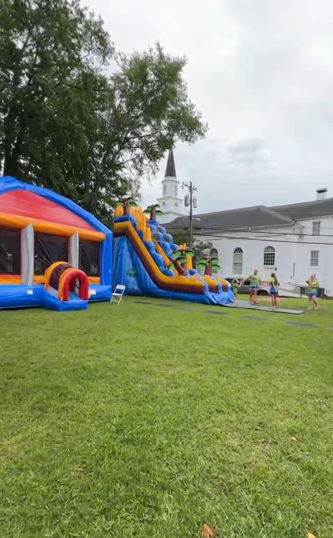 Inflatable bounce houses and slides on a grassy lawn at an outdoor event, with a white building in the background