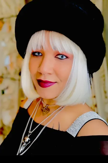 Portrait of a person with platinum bob hair, black hat, silver necklaces, and pink lipstick, smiling indoors