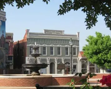 Historic gray mansion with a fountain, framed by trees and a brick wall in the foreground