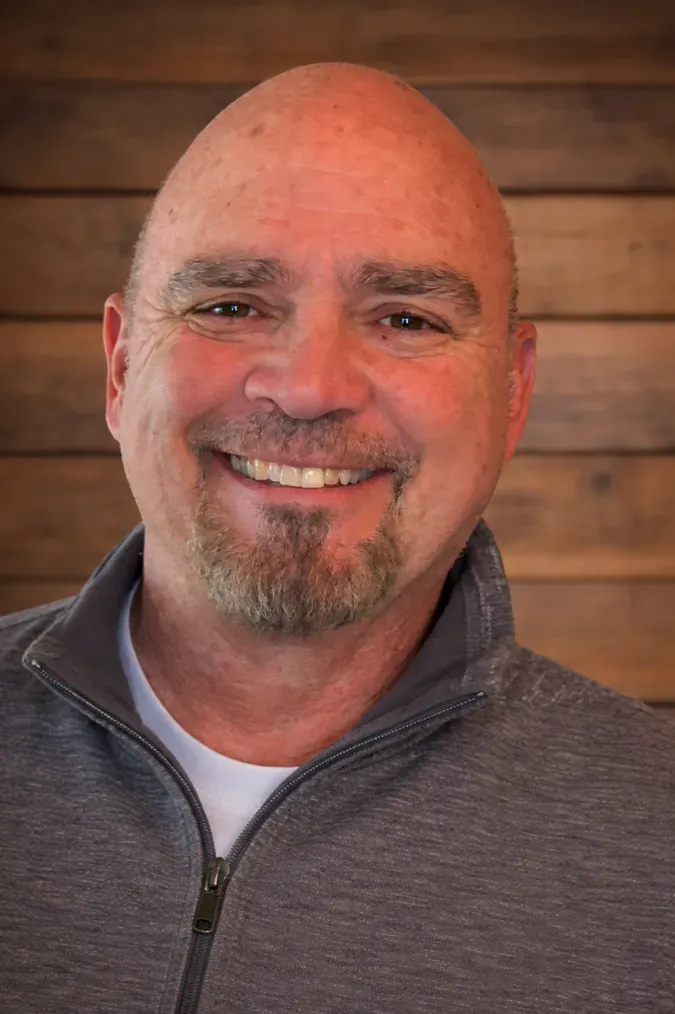 Smiling bald man with a goatee wearing a gray zip-up sweater against a wooden wall