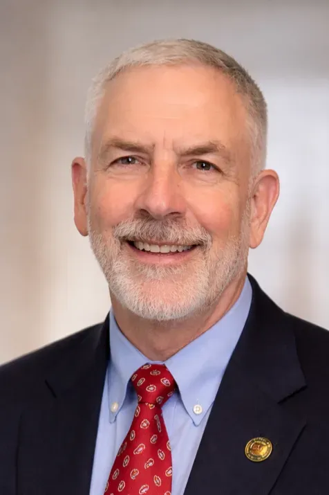 Smiling man in navy suit, light blue shirt, and red tie against a neutral background