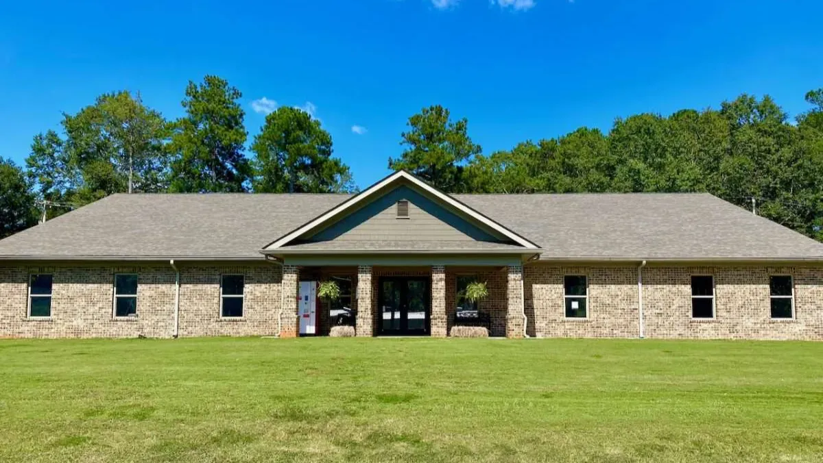 Single-story stone building with a central porch, green roof trim, and lawn under a blue sky.