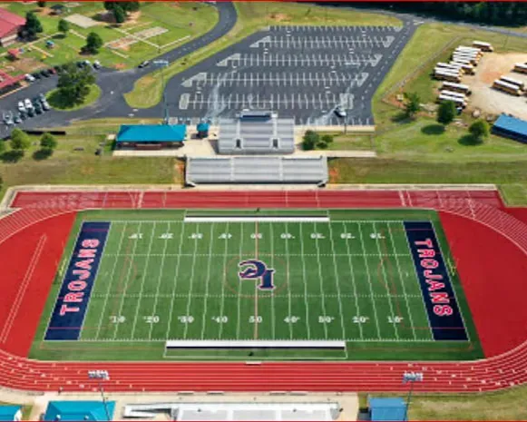 Aerial view of a football field with red track and “Trojans” end zones beside a large parking lot.