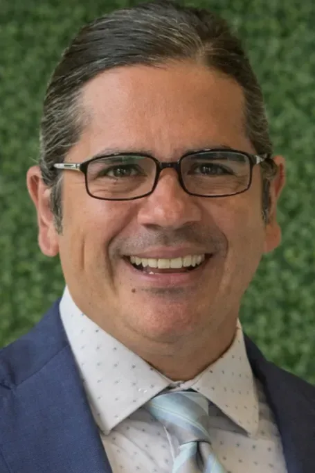 Smiling man in glasses and suit jacket, outdoors with blurred greenery background