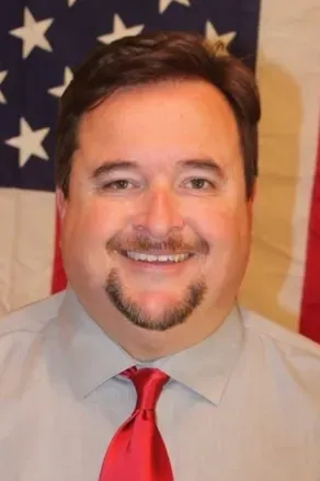 Man in gray shirt and red tie smiling in front of an American flag backdrop