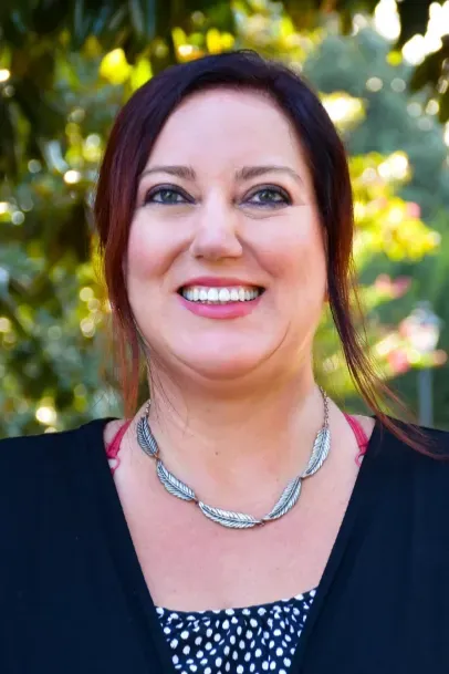 Smiling woman outdoors with dark hair, black top, and silver necklace against a blurred green background