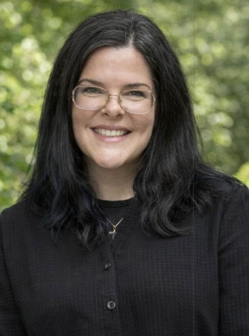 Smiling woman with glasses and dark hair with blue streaks, standing against a light brick wall