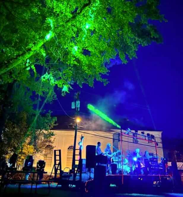 Outdoor nighttime concert with green lights, performers on stage, and a lit tree overhead