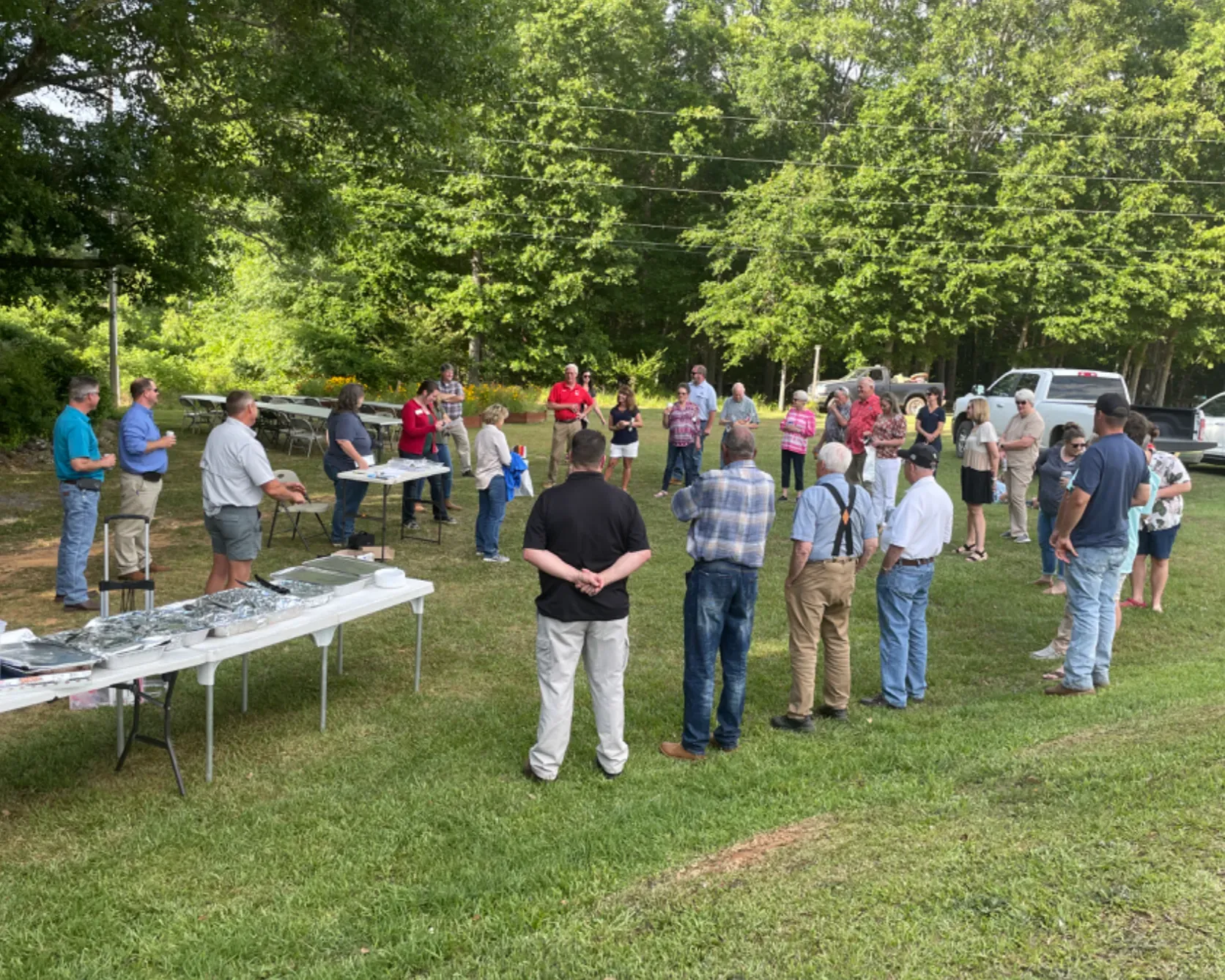 People gathered outdoors at tables in a grassy park, socializing near parked cars and trees.