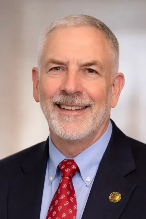 Portrait of a smiling man in a navy suit, light blue shirt, and red tie against a neutral background