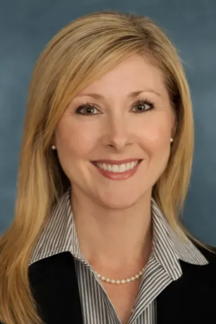 Smiling blonde woman in a black blazer and striped shirt against a blue studio background