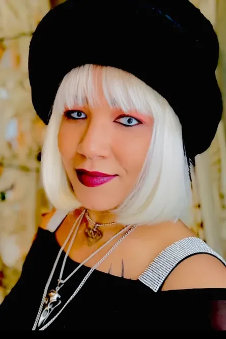 Portrait of a person with platinum bob hair, black hat, and silver chain necklace, wearing black and smiling.