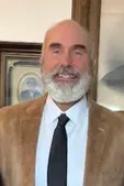 Smiling bald man with a gray beard wearing a tan suit, white shirt, and black tie indoors