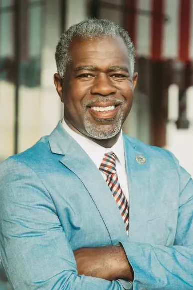 Smiling man in a light blue suit with arms crossed against a blurred indoor background