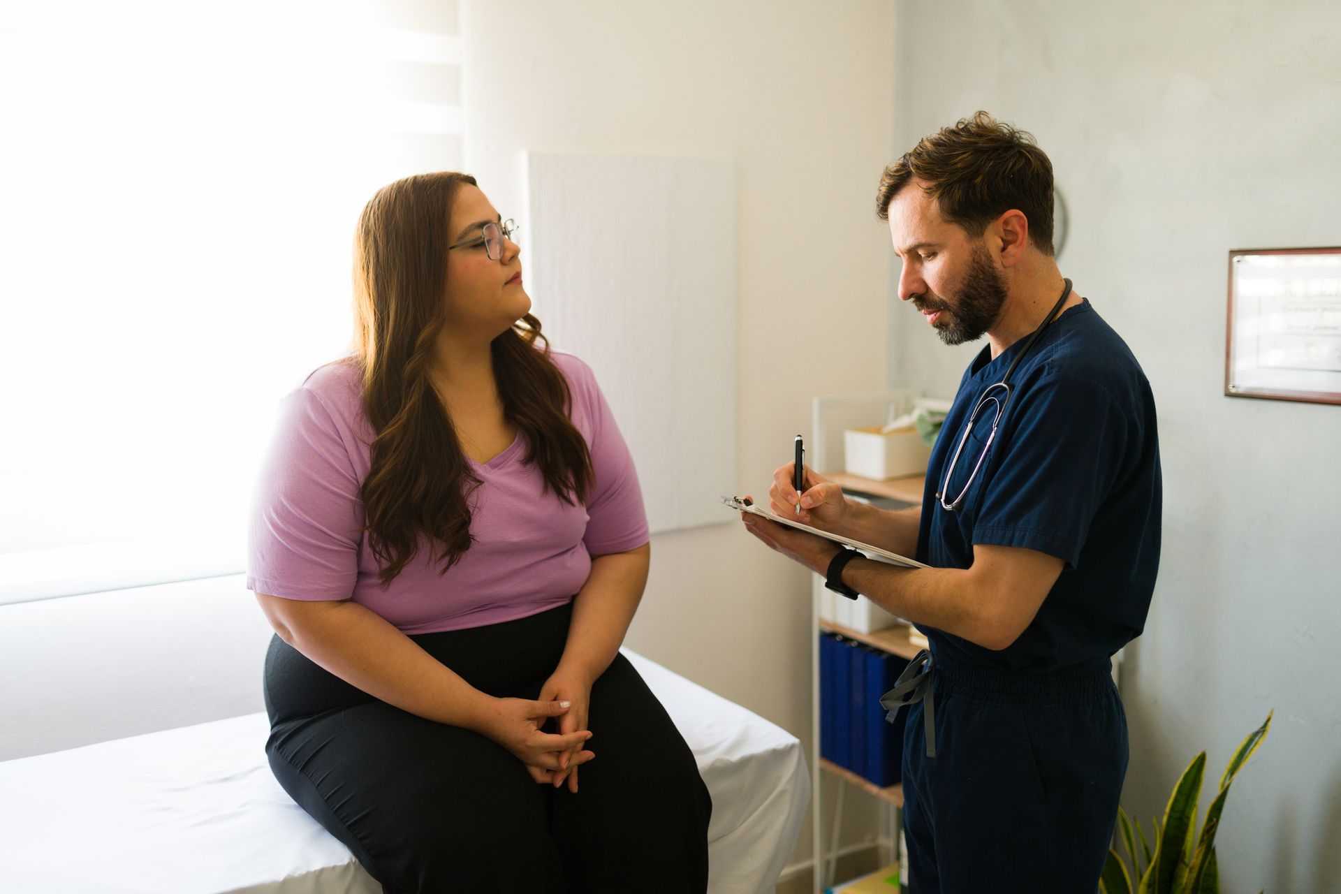 Doctor taking notes while speaking with a seated patient in a bright exam room