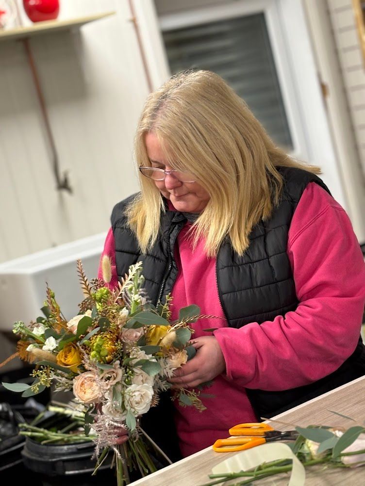 Woman arranging a bouquet, wearing pink sweater and black vest. Workshop setting with flowers and tools.