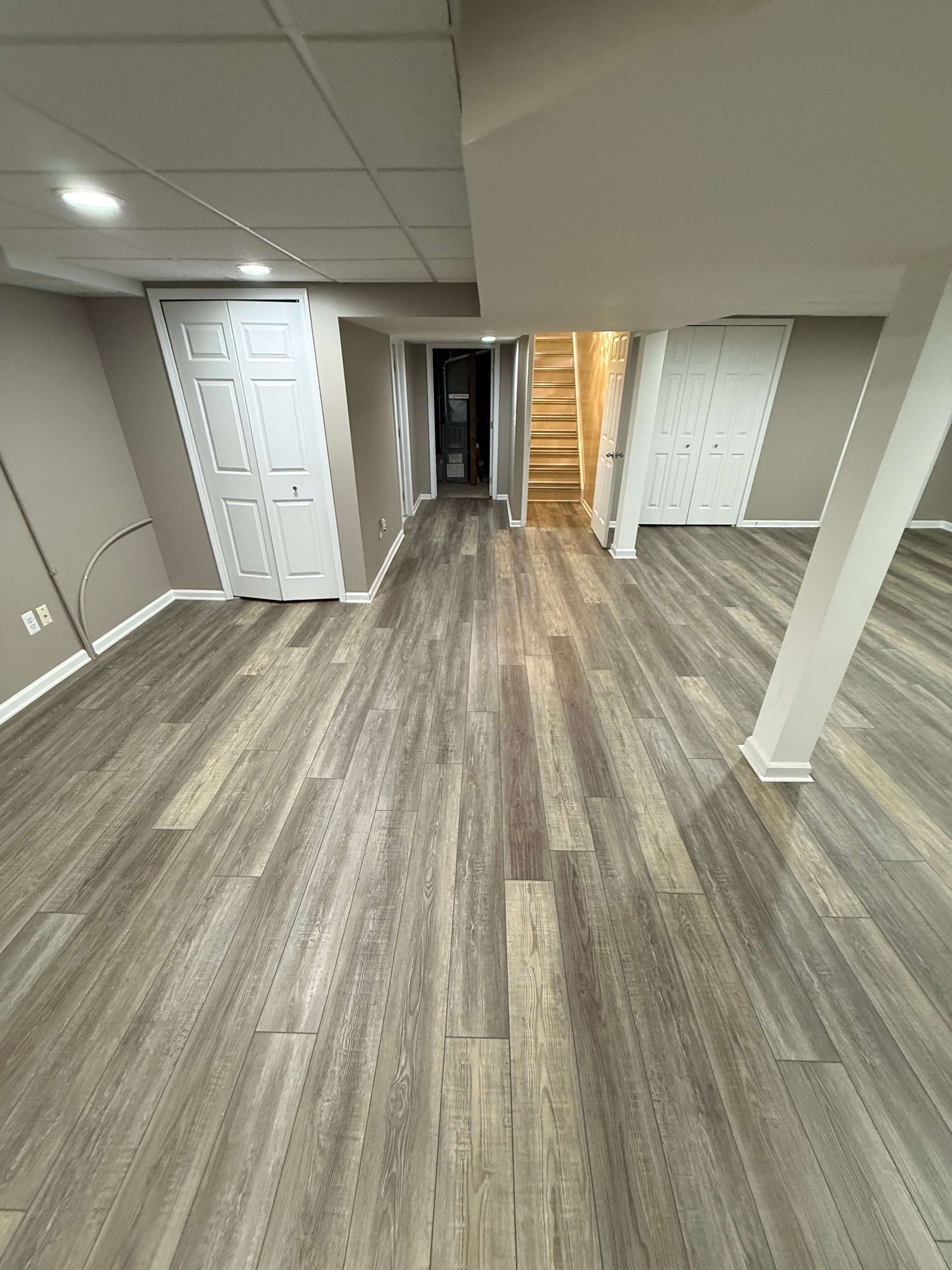 Empty basement room with gray wood floors, white doors, and stairs in the back.