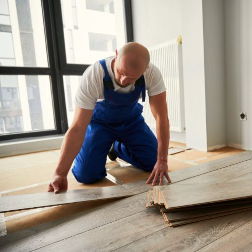 Worker installing wooden flooring in a bright room, kneeling beside stacked planks